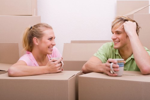 In a residential setting, a young woman and a young man are sitting among stacked cardboard moving boxes, engaging in a conversation and smiling. The woman, on the left, has blonde hair tied back and is wearing a pink top, while the man, on the right, has blonde hair and is dressed in a light green t-shirt, resting his head on his hand. A mug is placed in front of the man, and the woman holds a small object, possibly a phone or a remote. The scene appears to be inside a house, with a plain white wall in the background and natural lighting illuminating their faces. The boxes are made of brown cardboard with some visible creases and tape, indicative of a recent or ongoing move within or around Fulham, London, with the activity suggesting house relocation or packing preparation. The setting reflects typical household content associated with residential moves, with a focus on the emotional and social aspects of packing and planning for a house removal through services like those offered by Fulham Man with Van.