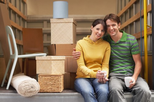 A young couple sitting on the floor of a residential storage unit or moving truck, surrounded by unpacked cardboard boxes of various sizes and textures, including woven and plain cardboard finishes. The woman is wearing a yellow hoodie and holding a mug, while the man is dressed in a green and purple striped polo shirt, also holding a mug, both smiling at the camera. Behind them, shelves are partially visible, with some storage items and a rolled-up rug placed to the side. A tall, light blue canister sits atop a stacked arrangement of boxes, positioned near the back of the space. The setting suggests a house moving or unpacking process within a furnished or partially furnished interior space in Fulham, London, with a neutral lighting atmosphere. The scene reflects typical household items involved in removals, emphasizing the process of packing and relocation. Fulham Man with Van services might facilitate such moves, supporting local residents in the SW6 postcode area.