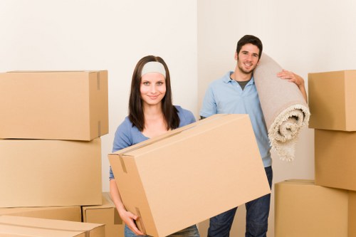 A young woman and a young man are inside a residential space, likely a house awaiting or in the process of moving. The woman, standing in the foreground, holds a large, rectangular cardboard box with a plain brown exterior, suggesting it is packed for a move. She has shoulder-length dark hair, wears a light grey headband, and is dressed in a blue long-sleeved top. The man, positioned slightly behind her to the right, is smiling and carries a rolled-up beige carpet or rug over his shoulder, gripping it with one hand. He has short dark hair, a neatly trimmed beard, and is dressed in a light blue shirt rolled up at the sleeves and dark trousers. Around them, several other cardboard boxes of different sizes are stacked or placed on the floor, some of which are closed while others are open, indicating an active packing or unpacking process. The background features plain white walls with no visible decorations or furniture, and the lighting appears to be natural and bright, providing clear visibility of all objects. This scene suggests an active house removal, consistent with local removals services in Fulham or nearby areas, with the company Fulham Man with Van likely involved in facilitating this move.