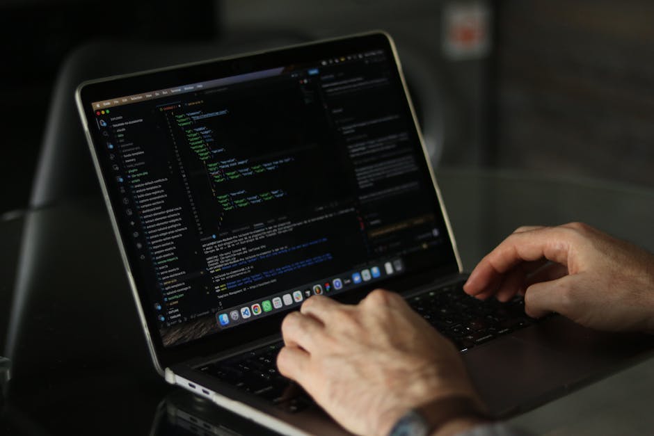 A close-up view of a person's hand wearing a wristwatch, typing on a laptop keyboard placed on a wooden surface. The laptop screen displays lines of code with colored syntax highlighting, suggesting e