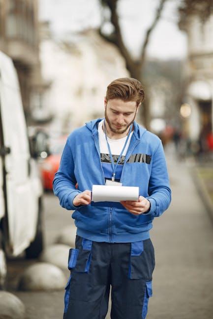A young male staff member wearing a blue uniform with darker blue work trousers stands outdoors on a city street during daytime, holding a clipboard or tablet device and reviewing documents or plans. 