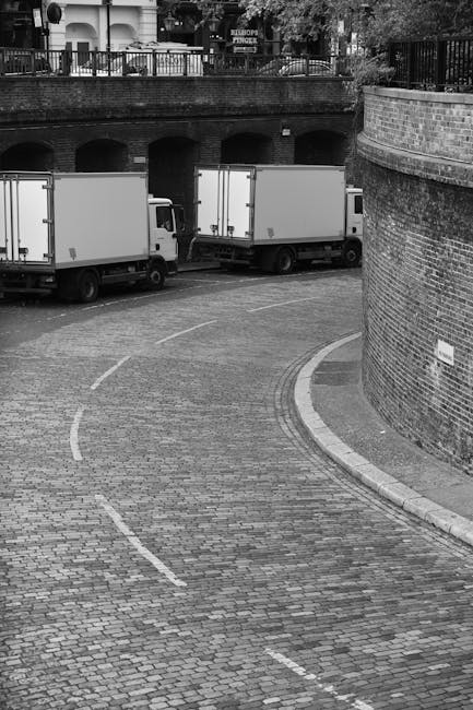 A black-and-white photograph showing a curved cobblestone driveway adjacent to a brick wall, with three large white removal trucks parked along the curb. The trucks are positioned parallel to the pave